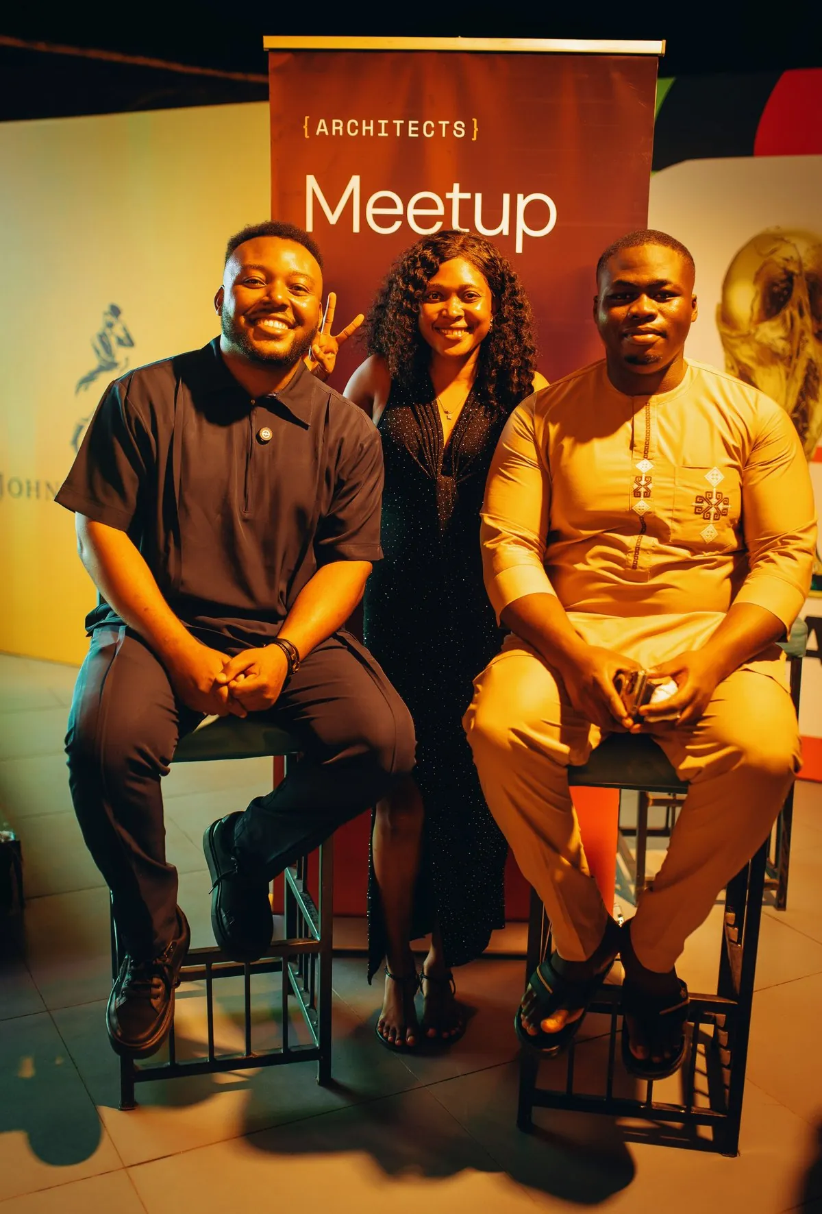 Two hosts seated on high stools either side of a speaker, beneath an {ARCHITECTS} Meetup banner during a community panel.